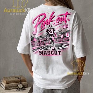 Puede incluir: Una camiseta blanca con un gr&aacute;fico en la espalda de un jugador de f&uacute;tbol americano con el n&uacute;mero 23 y el texto "YOUR NAME". El dise&ntilde;o incluye "Pink out." y "MASCOT" con cintas rosas y luces de estadio.