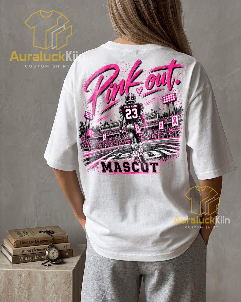 Puede incluir: Una camiseta blanca con un gr&aacute;fico en la espalda de un jugador de f&uacute;tbol americano con el n&uacute;mero 23 y el texto "YOUR NAME". El dise&ntilde;o incluye "Pink out." y "MASCOT" con cintas rosas y luces de estadio.