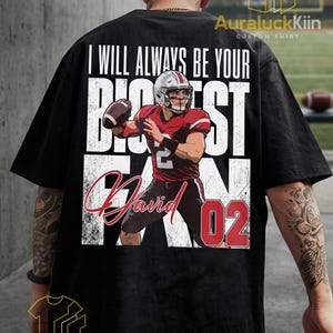 Puede incluir: Una camiseta negra con el gráfico de un jugador de fútbol americano con camiseta roja y casco sosteniendo un balón. El texto dice I WILL ALWAYS BE YOUR BIGGEST FAN, con el nombre David en letra roja y el número 02 en letras rojas.