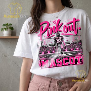Puede incluir: Una camiseta blanca con un estampado gr&aacute;fico de un jugador de f&uacute;tbol americano que lleva una camiseta con el n&uacute;mero 23 y el texto YOUR NAME en la espalda. El dise&ntilde;o incluye las palabras Pink out y MASCOT en letras rosas con lazos.
