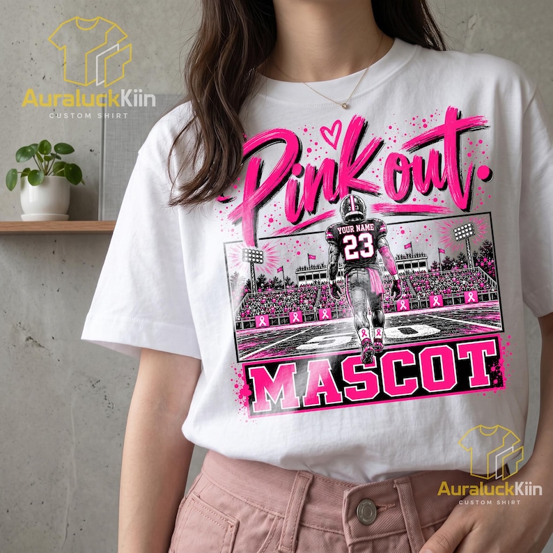 Puede incluir: Una camiseta blanca con un estampado gr&aacute;fico de un jugador de f&uacute;tbol americano que lleva una camiseta con el n&uacute;mero 23 y el texto YOUR NAME en la espalda. El dise&ntilde;o incluye las palabras Pink out y MASCOT en letras rosas con lazos.