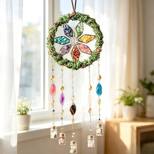 Può includere: Una decorazione da parete sospesa con una ghirlanda circolare di fogliame verde. All'interno della ghirlanda si trova una cornice a forma di stella riempita di frammenti di pietre preziose multicolori. Cinque catene pendenti hanno pietre.