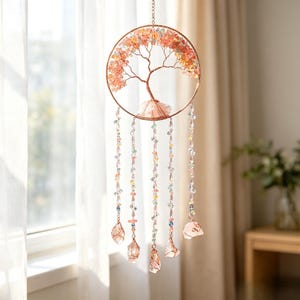May include: A circular wall hanging featuring a copper wire tree of life sculpture filled with small pink and light green crystal chips. Five dangling strands of multi-colored beads and clear crystals hang from the base, each ending in a raw crystal.