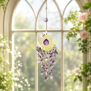 May include: A decorative suncatcher with a gold-colored sun and crescent moon design, holding a large purple gemstone. Strands of purple and clear crystals hang below. The suncatcher is suspended by a chain and has a tag that says "made LOVE".