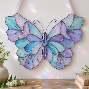May include: A decorative stained glass butterfly in shades of blue, purple, and lavender. The butterfly features a central jewel and is suspended by a lavender ribbon. The wings are made of multiple glass pieces, creating a mosaic effect.