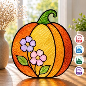 Puede incluir: Un atrapasoles de vidriera con forma de calabaza, con un cuerpo texturizado en naranja y amarillo y un tallo verde. La calabaza está decorada con dos flores de color morado claro y rosa con hojas verdes, delineadas con bordes de metal negro.