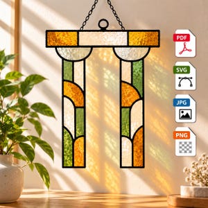 Könnte beinhalten: Ein Buntglas-Suncatcher mit einem architektonischen Bogendesign und zwei vertikalen Säulen. Das Stück besteht aus strukturierten bernsteinfarbenen, grünen und klaren Glasscheiben, die von schwarzen Metallprofilen gehalten und an einer Kette aufgehängt sind.