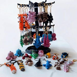 May include: A black rotating display stand holds numerous articulated animal keychains in various colors and designs, including octopuses, sharks, turtles, and fish. Additional loose keychains are arranged on the surface below the stand.