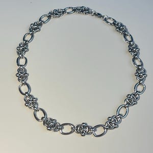 Op de afbeelding: Een zilverkleurige ketting met een afwisselend patroon van gladde, langwerpige ovale schakels en complexe, gegroepeerde cirkelvormige ringsegmenten. De metalen schakels zijn gepolijst en reflecterend en vormen een doorlopend cirkelvormig ontwerp.