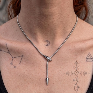 Könnte beinhalten: Eine silberfarbene Schlangenkette mit einem zentralen metallischen Kopfelement, das einen einzelnen klaren Stein enthält. Die Kette verläuft durch das Kopfteil und bildet einen Y-förmigen Anhänger, der in einer spitzen, konischen Metallform endet.