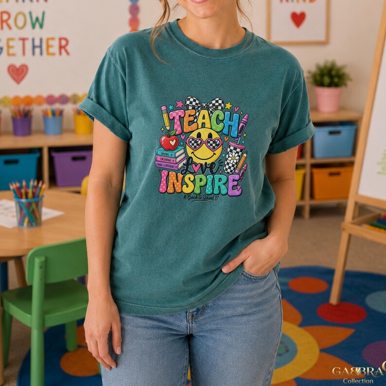 May include: A teal short-sleeve t-shirt featuring a graphic print with a yellow smiley face wearing checkered sunglasses, a bow, books, a pencil, and a cup. The text reads TEACH LOVE INSPIRE with the hashtag #BackToSchool in a colorful font.