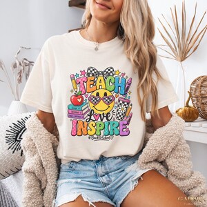 May include: A cream-colored short-sleeve t-shirt featuring a colorful graphic print with the text TEACH LOVE INSPIRE and #BacktoSchool. The design includes a smiley face with heart-shaped glasses, a stack of books, an apple, and a checkered bow.