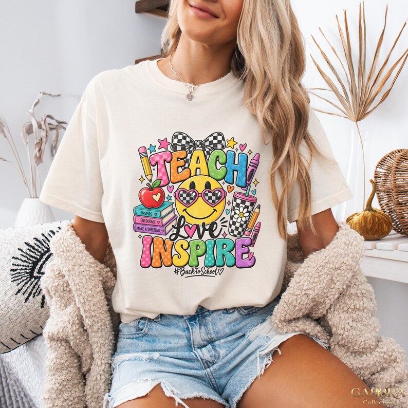 May include: A cream-colored short-sleeve t-shirt featuring a colorful graphic print with the text TEACH LOVE INSPIRE and #BacktoSchool. The design includes a smiley face with heart-shaped glasses, a stack of books, an apple, and a checkered bow.