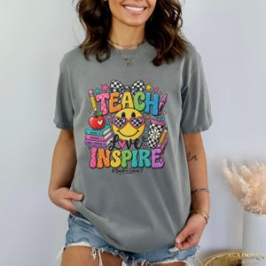 May include: A grey short-sleeve t-shirt featuring a colorful graphic print with the text TEACH and INSPIRE. The design includes a yellow smiley face wearing heart-shaped glasses, a stack of books, a red apple, and pencils in a checkered cup.