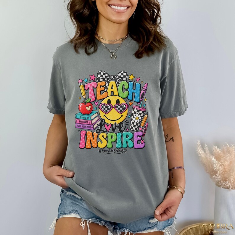 May include: A grey short-sleeve t-shirt featuring a colorful graphic print with the text TEACH and INSPIRE. The design includes a yellow smiley face wearing heart-shaped glasses, a stack of books, a red apple, and pencils in a checkered cup.