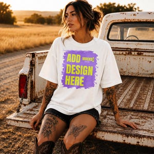 May include: A white short-sleeved t-shirt featuring a purple brushstroke graphic with the yellow text ADD DESIGN HERE and the small logo MOCKD. The shirt has a crew neckline and a loose fit, worn with black shorts and dark brown cowboy boots.
