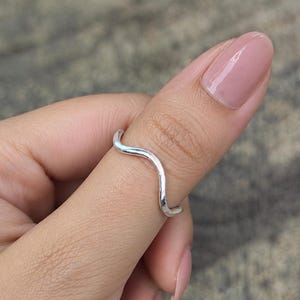 May include: A thin, silver-coloured ring with a single smooth, wave-like curve. The band has a polished, reflective metallic finish and maintains a consistent thickness throughout its design, encircling the thumb in a simple, continuous loop.