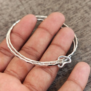 May include: A silver-toned bangle bracelet featuring a double-layered design with a hammered, textured surface. The bracelet has a circular shape and includes a small, heart-shaped loop detail at the closure point, creating a continuous band.