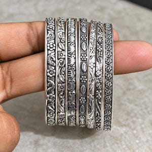 Può includere: Un set di sei bracciali rigidi in metallo color argento con intricati motivi in rilievo, tra cui disegni floreali, una fila di elefanti, forme geometriche e il simbolo Om, rifiniti con una texture ossidata scura per profondità.