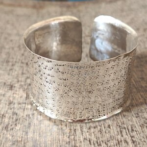 May include: A wide silver-colored cuff bracelet featuring an open back design. The exterior surface of the metal band is covered in a repeating, textured pattern of small, recessed geometric shapes that create a reflective, metallic finish.