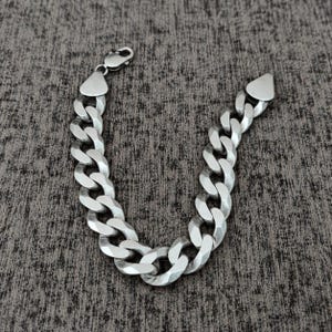 Può includere: Un bracciale a catena gourmette color argento con maglie spesse, intrecciate e sfaccettate. Il bracciale presenta una chiusura a moschettone sicura su un'estremità e un terminale in metallo piatto di forma triangolare sull'altra.