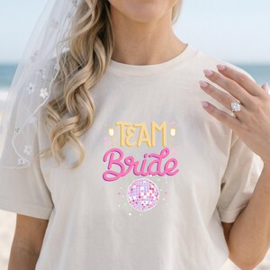 May include: A cream-colored short-sleeve t-shirt featuring the text "TEAM" in yellow and "Bride" in pink script. The design includes two yellow champagne flutes and a pink and white disco ball graphic with small colorful star accents.