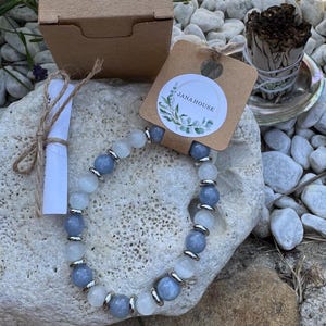 Könnte beinhalten: Ein Perlenarmband mit abwechselnd hellblauen und durchscheinend weißen runden Perlen, die durch kleine silberfarbene Metallabstandshalter getrennt sind. Das Armband ist an einem braunen Pappanhänger mit dem Text Jana House und einem Blattmotiv befestigt.