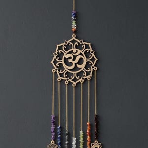 Op de afbeelding: Een wandhanger met een lasergesneden houten Om-symbool in een mandala-frame, opgehangen aan een goudkleurige ketting. Daaronder hangen zeven verticale kettingen, elk versierd met veelkleurige edelsteensplinters en kleine, gedetailleerde houten chakra-symbolen.