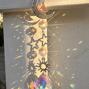 May include: A hanging suncatcher featuring a silver-toned crescent moon, a star, and a sun charm connected by metal links. The ornament includes clear, multifaceted glass crystals that refract sunlight, casting vibrant rainbow light patterns.