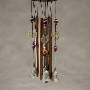 Pode incluir: Um carrilhão de vento com um topo circular de madeira vermelha, cinco tubos metálicos cor de cobre e três pequenos sinos em tom de latão. A estrutura é decorada com contas de madeira castanho-escuras e três moedas circulares de latão.