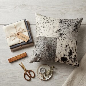 Könnte beinhalten: Ein quadratisches Kissen mit einem Patchwork-Design aus schwarz-weiß-grauem Tierfellmuster. Daneben liegen gefaltete Stoffquadrate, die mit einer Schnur und einem HANDMADE-Etikett versehen sind, ein Holzlineal, eine Metallschere und eine Kerze.