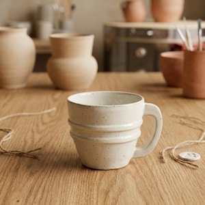 May include: A speckled off-white ceramic mug with a wide, rounded handle and two horizontal ridges encircling the body. The mug features a smooth, glazed finish with subtle dark flecks throughout the surface, resting on a wooden surface.