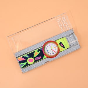 Könnte beinhalten: Eine Swatch-Armbanduhr mit rundem Gehäuse, roter Lünette und weißem Zifferblatt mit bunten Markierungen. Das Textilarmband hat eine dunkelblaue Basis mit neonpinken, gelben und grünen abstrakten Blumenmustern. Sie befindet sich in einer transparenten Kunststoffhülle.