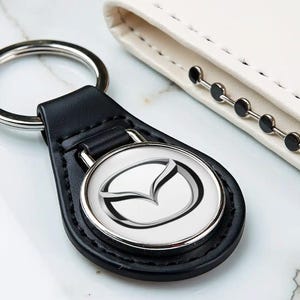 Può includere: Un portachiavi in pelle nera con un emblema metallico circolare che presenta il logo Mazda in argento e nero. Il portachiavi include un anello diviso in argento lucido per le chiavi ed è fissato a una linguetta in pelle nera con cuciture.