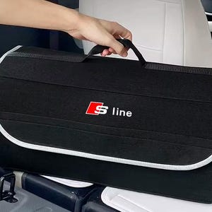 Peut inclure: Un organisateur de coffre de voiture rectangulaire noir doté d'une poignée supérieure et d'un rabat avec une bordure gris clair. L'avant présente un logo carré rouge avec un S blanc et le texte line en blanc, formant l'emblème S line.