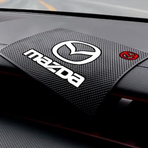 Könnte beinhalten: Eine schwarze, rechteckige, rutschfeste Armaturenbrettmatte mit strukturierter Oberfläche, die in der Mitte das weiße Mazda-Logo und den Schriftzug sowie rechts ein kleines, kreisförmiges, rot-weißes Mazda-Emblem-Symbol aufweist.