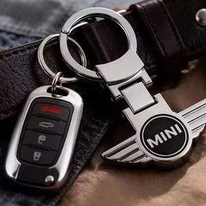 Op de afbeelding: Een zilverkleurige metalen sleutelhanger met het MINI-logo en gestileerde vleugels, bevestigd aan een ring. Daarnaast ligt een zwarte autosleutel met vier knoppen en een rood indicatielampje, afgewerkt met een gepolijste zilveren rand.
