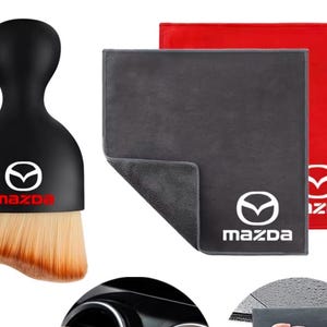 Puede incluir: Un cepillo negro para limpieza de interiores de coche con mango redondeado y el logotipo de Mazda, acompañado de un paño de microfibra gris y otro rojo, cada uno con el logotipo blanco de Mazda impreso en el centro de la tela.