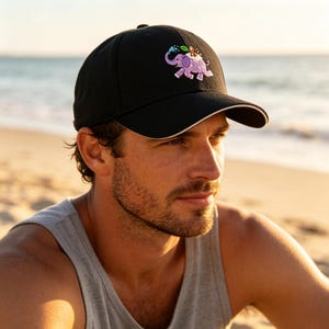 May include: A black baseball cap featuring an embroidered graphic of a purple elephant with orange spots spraying water from its trunk, with a small brown monkey sitting on its back. The cap has a structured crown and a curved brim with white trim.