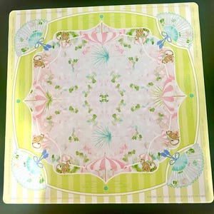 May include: A square scarf featuring a light green and white striped border with a central pattern of pink and white striped umbrellas, light blue folding fans, and small brown monkeys. The design includes repeating text elements.