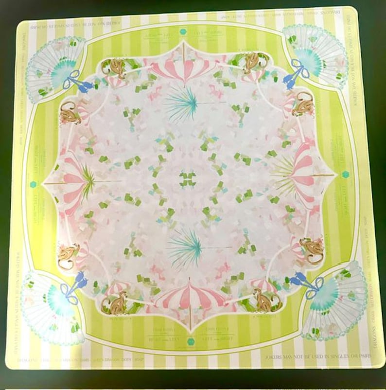May include: A square scarf featuring a light green and white striped border with a central pattern of pink and white striped umbrellas, light blue folding fans, and small brown monkeys. The design includes repeating text elements.