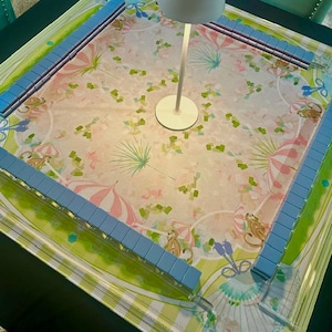May include: A square mahjong table mat featuring a light pink and green pattern with illustrations of monkeys and striped umbrellas. The mat is bordered by four light blue tile racks arranged in a square, with a white lamp centred on the surface.