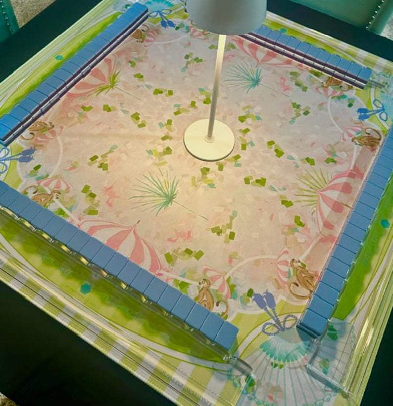 May include: A square mahjong table mat featuring a light pink and green pattern with illustrations of monkeys and striped umbrellas. The mat is bordered by four light blue tile racks arranged in a square, with a white lamp centred on the surface.