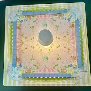 May include: A square board game set featuring a central light-coloured circular piece surrounded by four clear acrylic racks holding light blue tiles. The board surface displays a repeating pattern of pink umbrellas, green palm leaves, and small tan monkeys.