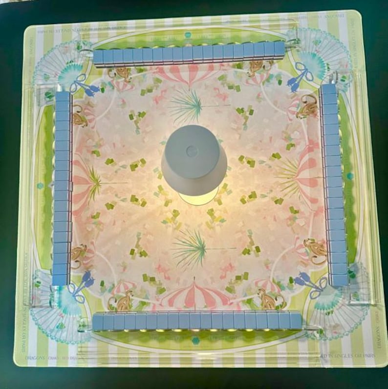 May include: A square board game set featuring a central light-coloured circular piece surrounded by four clear acrylic racks holding light blue tiles. The board surface displays a repeating pattern of pink umbrellas, green palm leaves, and small tan monkeys.