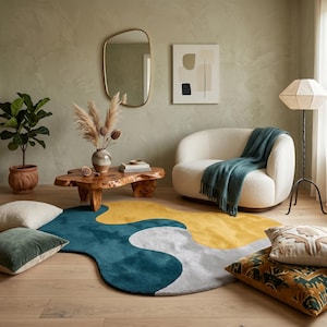 May include: An irregular, organic-shaped area rug featuring large, flowing color blocks of deep teal, mustard yellow, and light gray. The rug is placed on a light wood floor, surrounded by a cream-colored armchair, a wooden coffee table, and pillows.