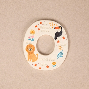 May include: A circular off-white ceramic plaque featuring a hand-painted lion, toucan, sun, and flowers. The text reads 16 February 2026, 51 CM, 3.4 KG, and I LOVE YOU baby. The design includes small orange stars and dots around the central opening.