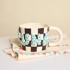 May include: A ceramic mug featuring a black and white checkered pattern. The word LUNA is written in raised, light blue, polka-dotted letters across the front. The mug has a curved handle and a smooth, glossy finish on the exterior surface.