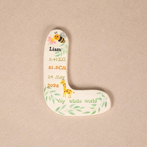 May include: A cream-colored, L-shaped ceramic plaque featuring a hand-painted bee, a pink flower, and a yellow giraffe. The text reads: Liam, 3.41 KG, 51.3 CM, 24 May 2026, and My whole world, surrounded by green leaf illustrations.