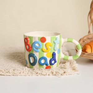 May include: A ceramic mug featuring light green vertical stripes and colorful polka dots. The mug has raised, three-dimensional letters in various colors spelling out the words "best dad" on the front. It includes a matching striped handle.